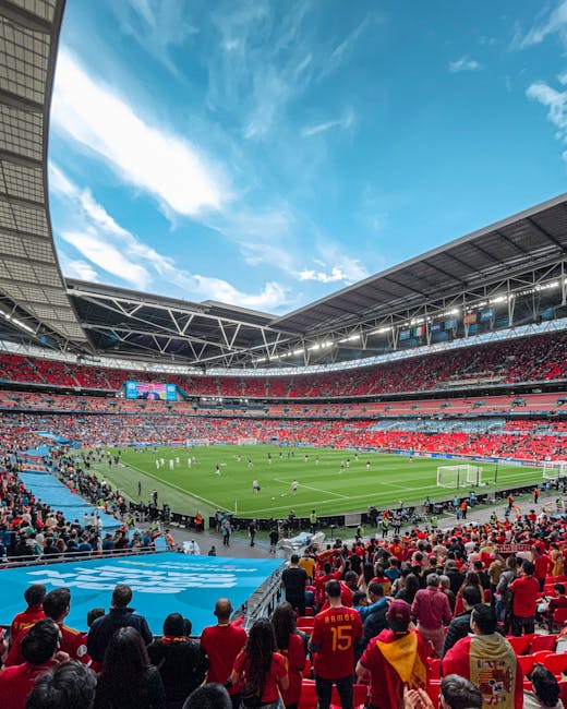 A wide view of Wembley Stadium's interior showing the tiered red seating areas and the lush green grass playing field with white boundary markings. The seating surrounds the field on all sides, with t